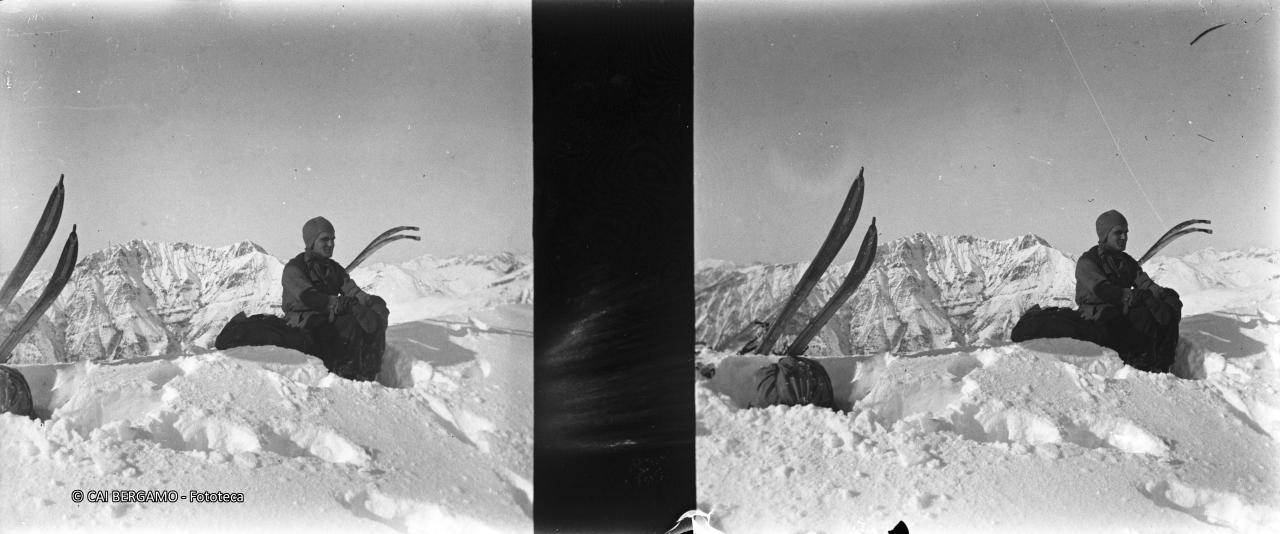 Skiatore con ski e zaino a riposo, sotto lo sguardo della retrostante cima di Menna