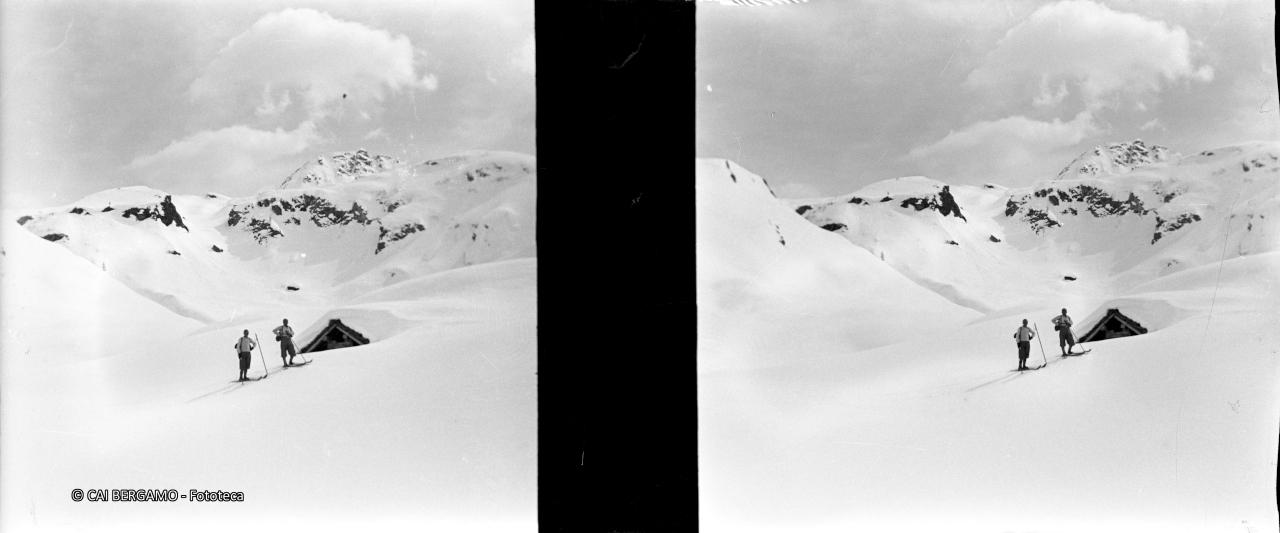 Due skiatori davanti a baita coperta di neve, sotto lo sguardo del Madonnino