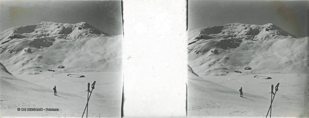 Skialpinista con bastoncini in primo piano e sguardo sulla cima  e sulle baite sottostanti coperte di neve